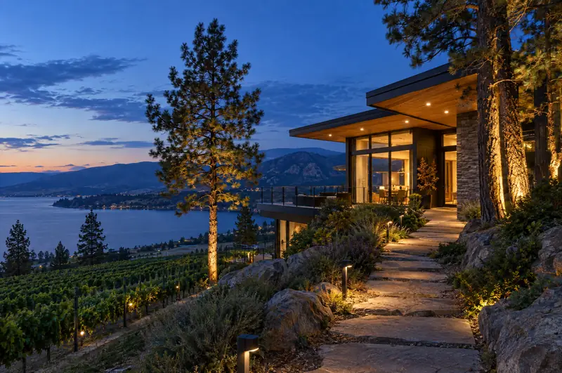 Lit stone path and uplit pine trees outside a hillside home at dusk